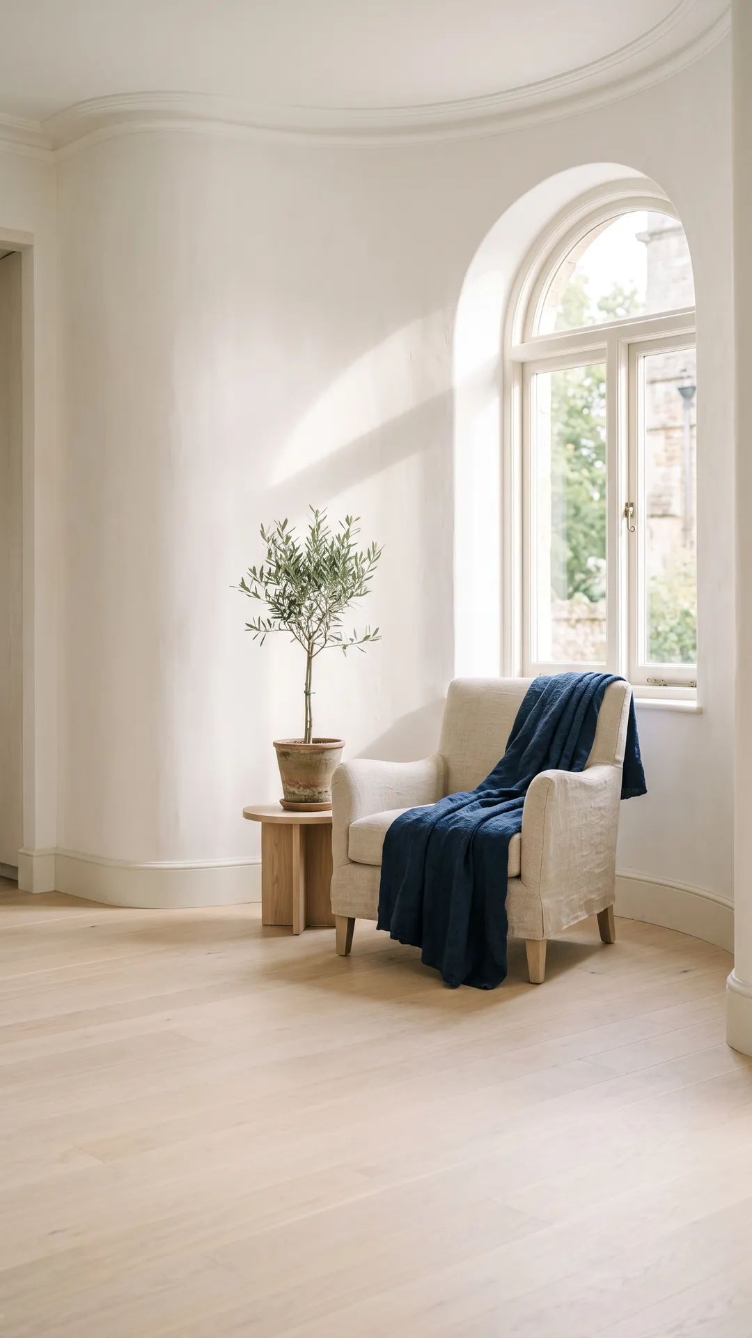 A linen armchair beside an arched window in a sunlit room, with an olive tree beside it.