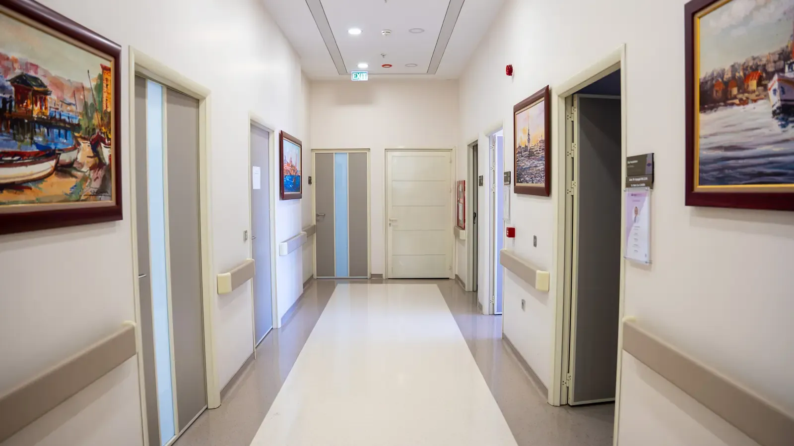 A daylit clinic corridor with pale walls, handrails and framed paintings.