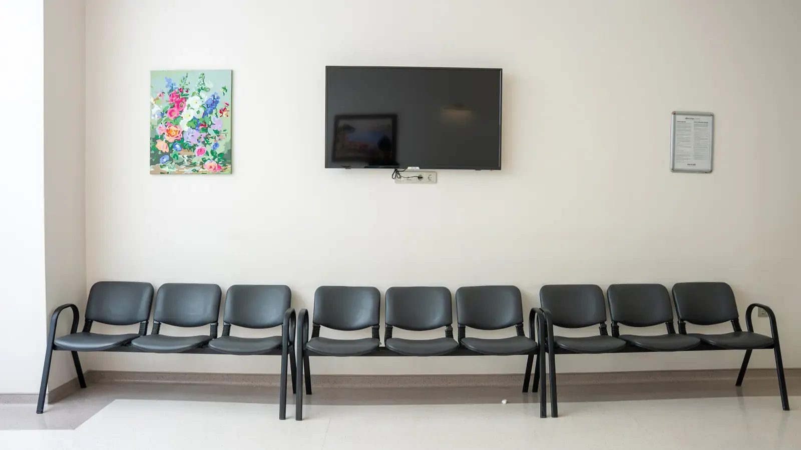 A row of seating along a pale wall beneath a wall-mounted screen and a framed painting.