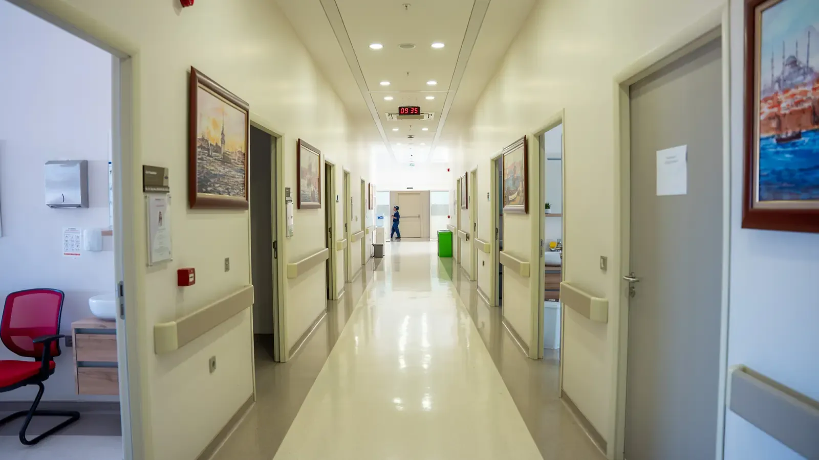 A daylit clinic corridor lined with doors and framed paintings, a staff member at the far end.